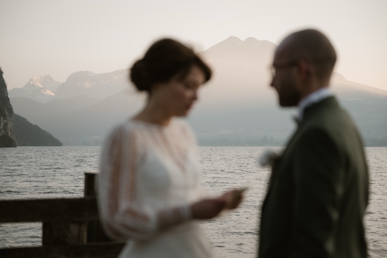 Out-of-focus portrait of bride and groom facing each other with Lake Annecy and mountains behind