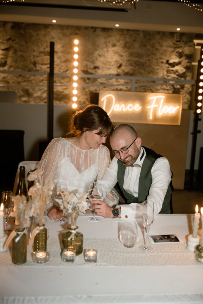 Bride and groom laughing together at their sweetheart table in front of a Dance Floor neon sign